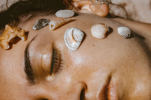 Woman posing with seashells on her face.\n\nWoman with seashells on her face daydreaming on the beach.
