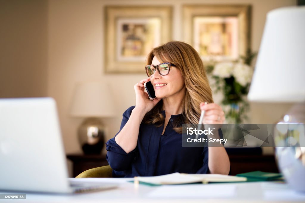 Happy businesswoman using mobile and laptop while working at home Shot happy businesswoman sitting at desk behind her laptop and talking with somebody on her mobile phone while working from home. Home office. Using Phone Stock Photo