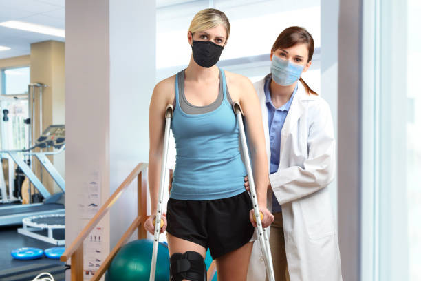 Physical Therapist Helping Patient With Use Of Crutches A female physical therapist helps a young athletic female patient who is wearing a knee brace with her crutches as they both look directly at the camera in a brightly lit physical therapy clinic. The therapist and the patient wear protective masks to prevent the spread of COVID-19 and other infectious diseases. knee brace stock pictures, royalty-free photos & images