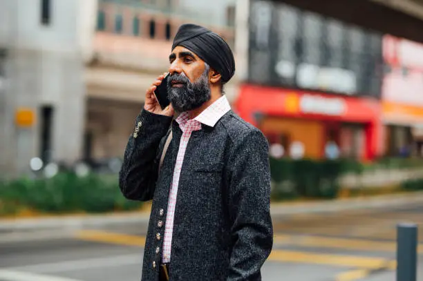 Portrait of Indian Sikh business man wearing business suit and black turban talking with smartphone in the city street.