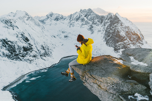 Woman sitting on cliff and using smartphone on Lofoten island in snow
