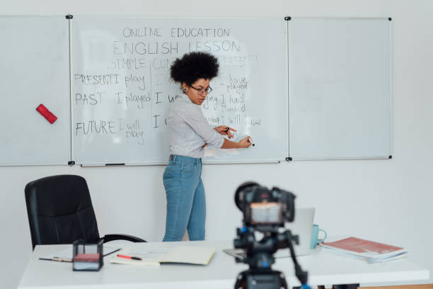 Young afro american woman standing near whiteboard, looking at web camera and explaining rules of English grammar, recording English lesson at home