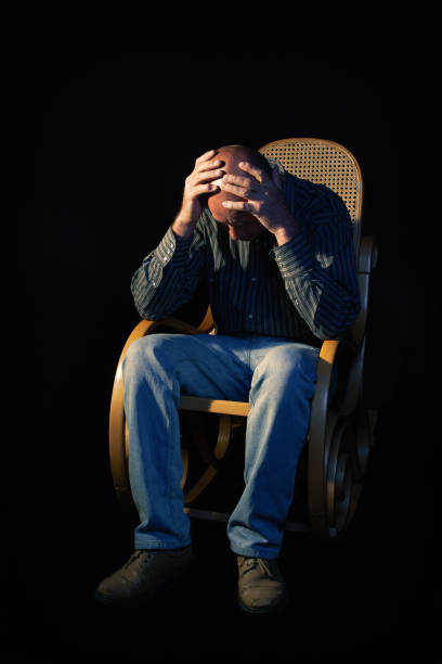 low key portrait of unemployed man thinking at the daily hardships sitting in a wooden arm rock chair isolated on black