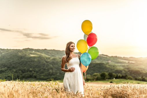 Beautiful young pregnant woman standing on a wheat field with balloons.