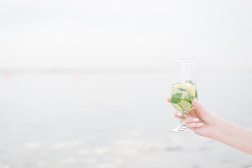 The hand of a young girl holds out a cocktail. Summer fruit drink in a glass with lime, rum and soda. Cold alcoholic mojito. The concept of relaxation, party and good mood. Copy space