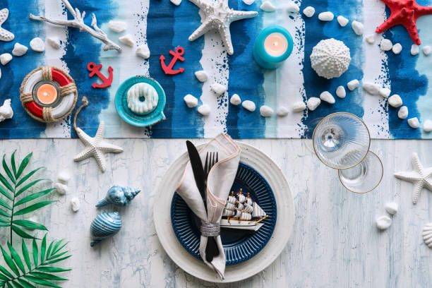 istock Columbus Day lunch table setting with nautical sea decorations on blue stripy runner, classic blue white plates, utensils on white rustic table. Top view, casual modern lunch, decorative sailing ship. 1259226210