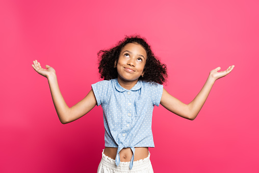 smiling cute curly african american kid showing shrug gesture isolated on pink