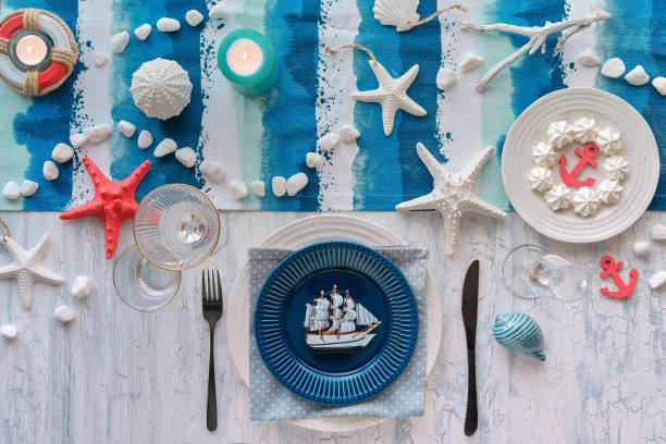istock Columbus Day lunch table setting with nautical sea decorations on blue stripy runner, classic blue white plates, utensils on rustic table. Top view, casual modern lunch setting. 1259227265