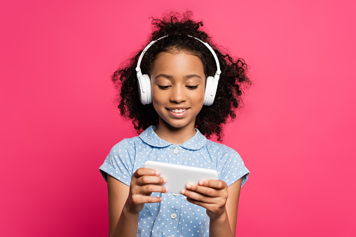 smiling curly african american kid in headphones using smartphone isolated on pink