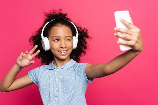 selective focus of smiling curly african american kid in headphones showing victory sign while taking selfie on smartphone isolated on pink