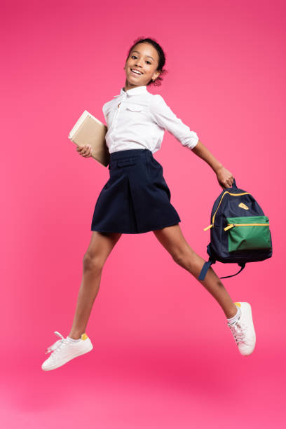 full length view of smiling african american schoolgirl jumping with backpack and books on pink background full length view of smiling african american schoolgirl jumping with backpack and books on pink background schoolgirl stock pictures, royalty-free photos & images