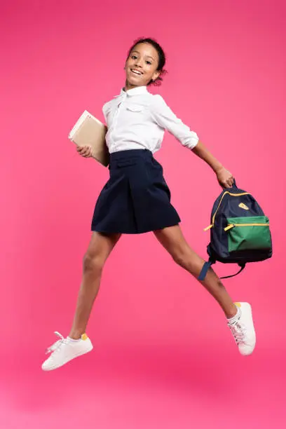 full length view of smiling african american schoolgirl jumping with backpack and books on pink background