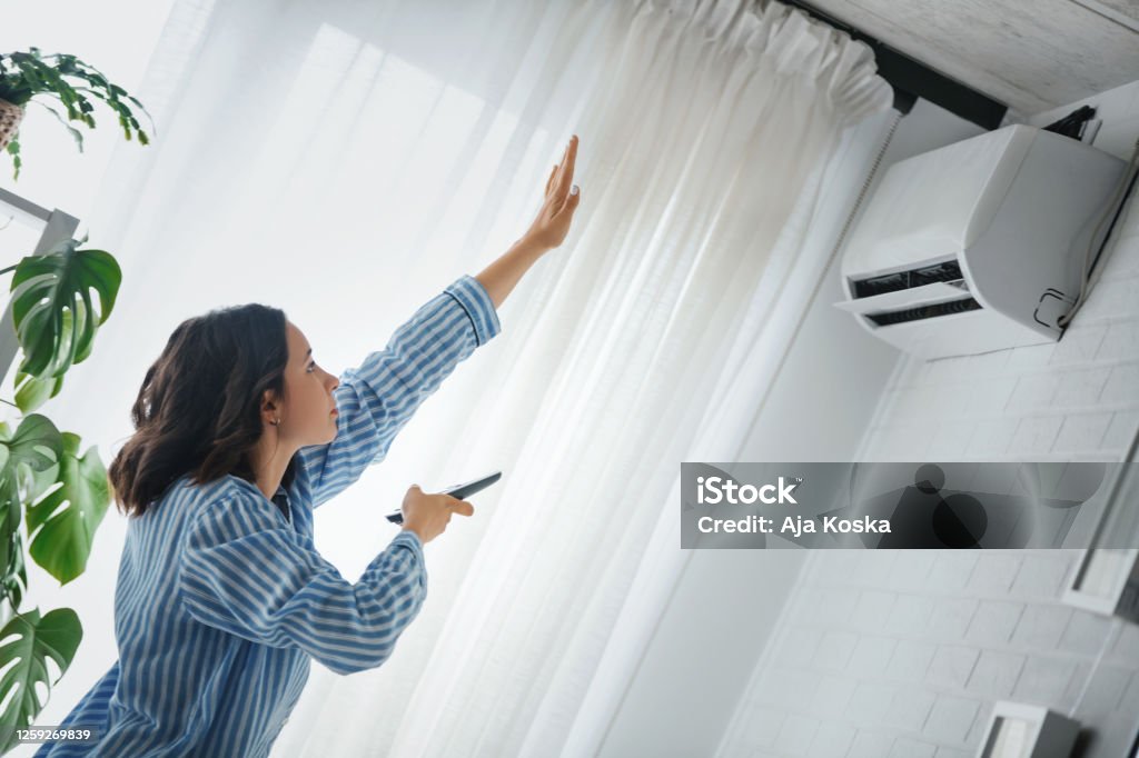 Turning on the air conditioner. Woman is checking to see if the air conditioner is cooling. She is holding the remote to the air conditioner and raised her hand to check temperature. Air Conditioner Stock Photo