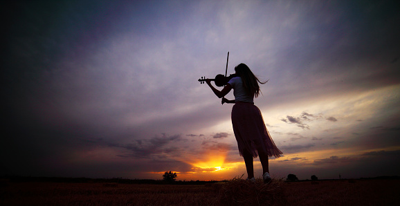 Romantic young woman with loose hair playing the violin in a field at sunset after harvest. Square sheaves of hay in the field. Violin training