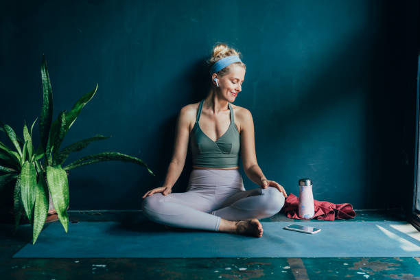 улыбаясь блондинка женщина с беспроводными наушниками, используя свой смартфон перед ее домашней тренировки - stretching yoga exercise mat women стоковые фото и изображения