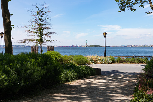 An empty path lined with green trees at The Battery Park with a view of the New York Harbor and the Statue of Liberty in New York City during the summer