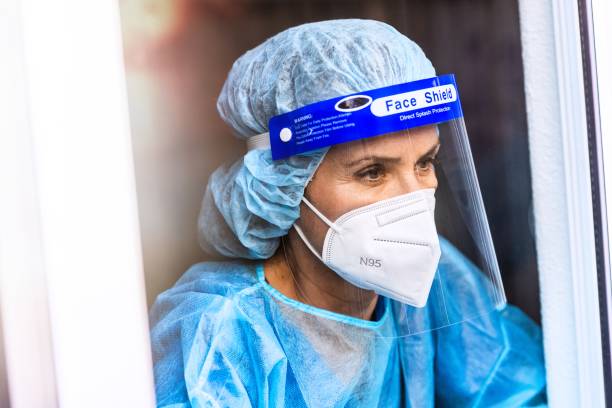 Tired, overworked, exhausted health care worker wearing a mask and a face shield posing looking away through a window