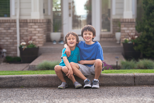 Two ethnic brothers (5 and 9) sit on a sidewalk in a residential neighborhood. They're smiling at the camera and have their arms around each other. There is a house in the background.