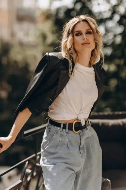 Photo of Stylish fashionable blonde woman with smoky eye makeup, in jeans, white t-shirt and black leather jacket on the balcony in the city. Spring autumn fashion concept. Soft selective focus.