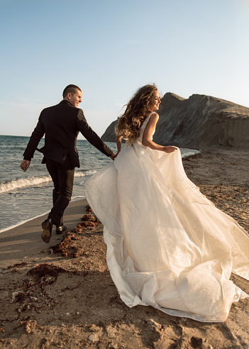 Happy and beautiful wedding couple run along the beach near the coastline holding hands against the background of rocks in the sea. A beautiful and smiling bride runs along the sand with a luxurious flowing hem of her dress.