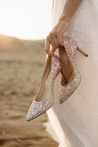 Close up photo of shiny luxurious high-heeled shoes with a golden sheen in the bride's hands in the sunny day.