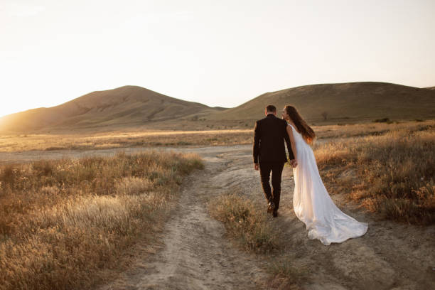 Bride and groom holding hands walk along a path in the field under the sunset mountains in a golden light. Wedding couple holding hands walks along a path in the field under the sunset mountains in a golden light. Bride with luxury flowing hem walks in safari holding hand of her lover and smiling. desert safari stock pictures, royalty-free photos & images