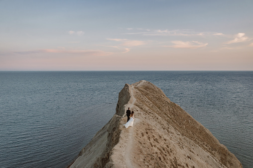 Bride and groom stand on top of a rock with a steep cliff into the ocean. The wedding dress flutters in the wind. The lovers stand in the distance and look at the horizon with the ocean and clouds.