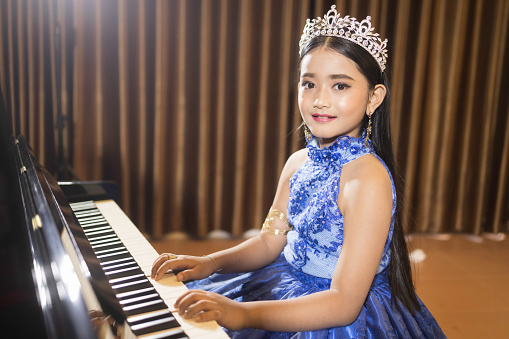 A portrait of a cute Asian girl, makeup, wearing a blue evening dress and a crown, playing the piano in the room.