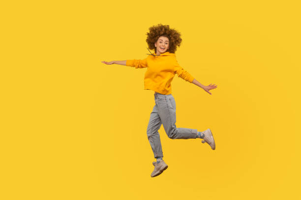 Portrait of enthusiastic lively energetic girl in urban style hoodie and jeans flying walking in air Portrait of enthusiastic lively energetic curly-haired girl in urban style hoodie and jeans flying walking in air, enjoying happy life, feeling inspired. studio shot isolated on yellow background air jump stock pictures, royalty-free photos & images