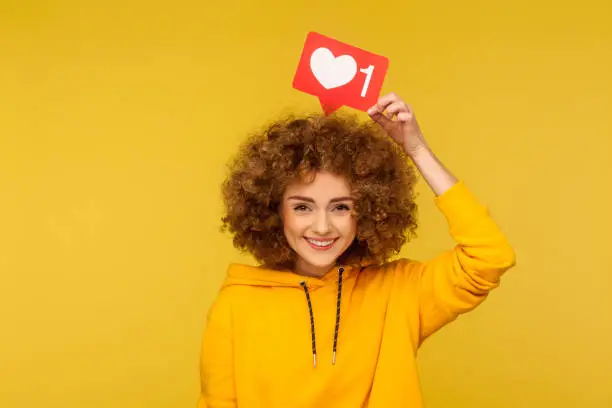 Photo of Internet blogging. Portrait of happy smiling curly-haired young woman in urban style hoodie holding heart like icon