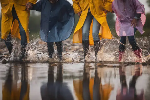 Unrecognizable family in raincoats having fun while stepping into big puddle in nature. Copy space.