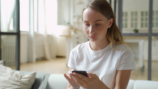 istock Young smiling woman using mobile phone while sitting on sofa at home 1259829687