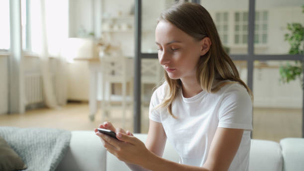 istock Young smiling woman using mobile phone while sitting on sofa at home 1259832271