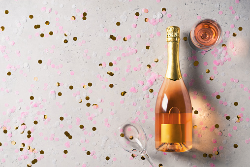 Bottle and glasses with rose sparkling wine on grey background with confetti, top view, copy space