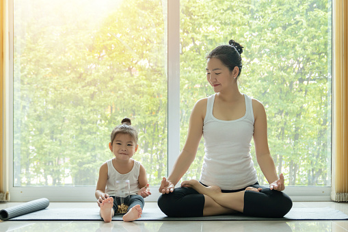 Asian mother practiced yoga by sitting in lotus position at home with a daughter sitting beside and enjoy eating snack with smiling face while workout at home. mom and kid doing activity at home
