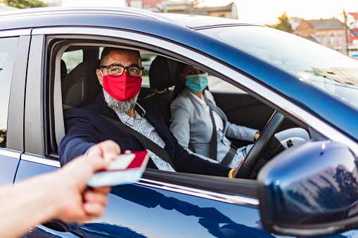 Photo of mature handsome man wearing glasses giving passport to staff at check point while sitting in his car with protective mask with his wife.