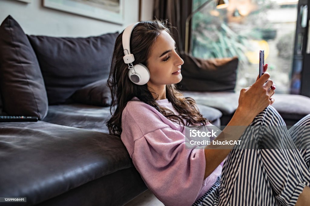 Young woman enjoying at home Photo of a young woman sitting on the floor in the living room, using smart phone and listening music Podcasting Stock Photo