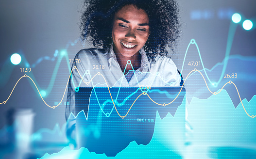 Cheerful young African American woman using laptop at blurry table with double exposure of financial graph. Concept of trading and financial success. Toned image