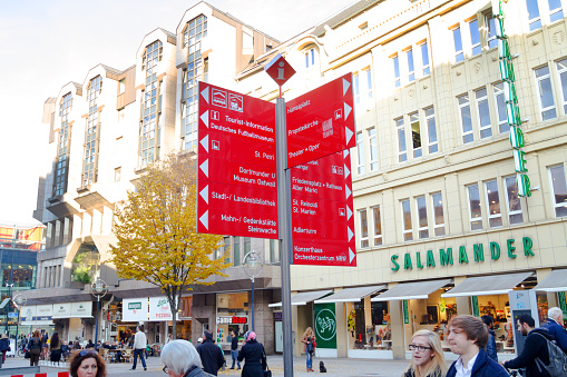 Pedestrian zone Westenhellweg and directions signs in Dortmund city - autumn season shop in main city street. In center are many direction signs as red signs with text and arrow. People are walking around. In right background is Salamander footwear sore