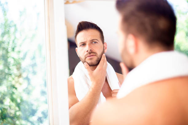 istock Handsome young man standing in front of bathroom mirror and looking his face 1259933472