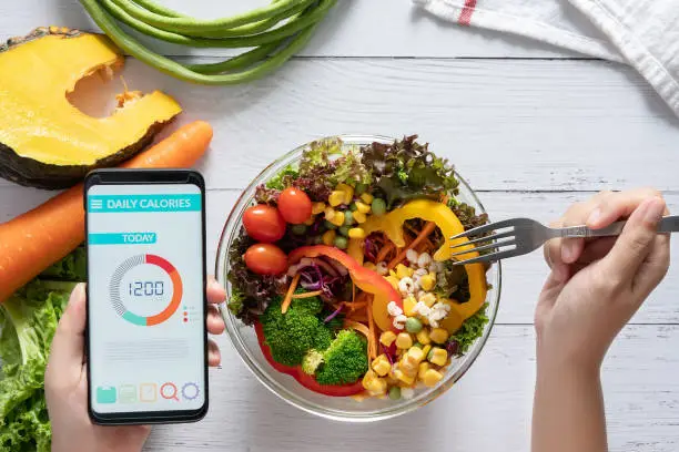Photo of Calories counting , diet , food control and weight loss concept. Calorie counter application on smartphone screen at dining table with salad, fruit juice, bread and fresh vegetable. healthy eating