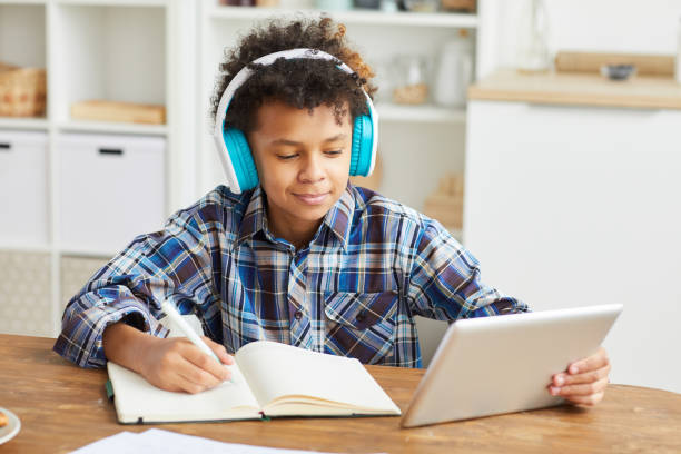 Boy studying at home African boy in headphones using digital tablet and making notes in his notebook while sitting at the table at home home schooling homework computer learning stock pictures, royalty-free photos & images