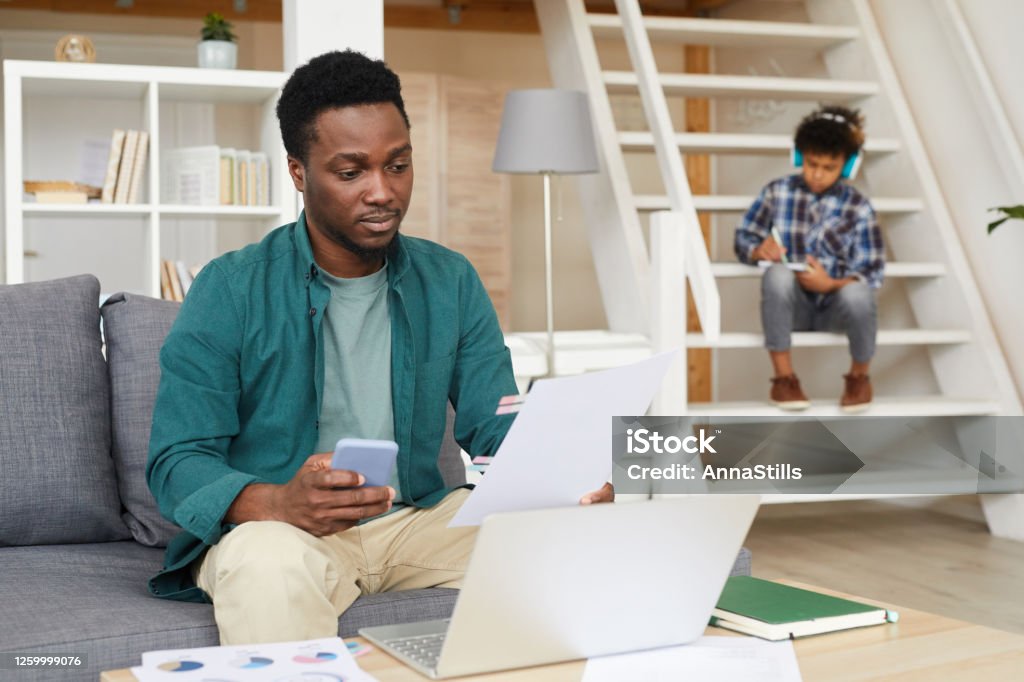 Working and studying online African young father sitting on sofa using laptop and examining documents with his son sitting in the background and studying online Child Stock Photo