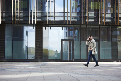 Wide angle portrait of elegant businessman wearing trenchcoat walking across shot while speaking by smartphone, glass urban building in background