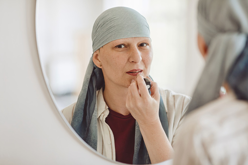 Minimal reflection portrait of mature bald woman putting on makeup and lipstick while looking in mirror at home, embracing beauty, alopecia and cancer awareness, copy space