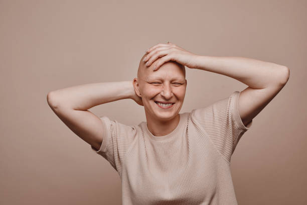 Carefree Bald Woman in Studio Warm-toned waist up portrait of carefree bald woman touching shaved head and smiling while posing against minimal beige background in studio, alopecia and cancer awareness, copy space balding photos stock pictures, royalty-free photos & images