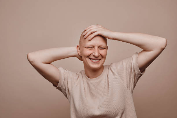 Carefree Bald Woman in Studio Warm-toned waist up portrait of carefree bald woman touching shaved head and smiling while posing against minimal beige background in studio, alopecia and cancer awareness, copy space balding stock pictures, royalty-free photos & images