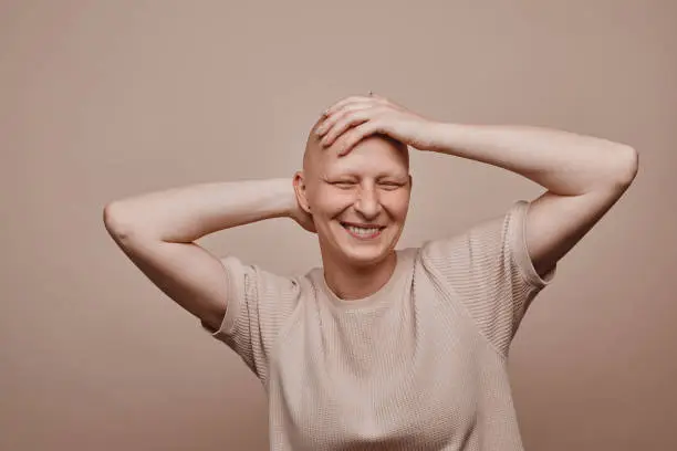 Warm-toned waist up portrait of carefree bald woman touching shaved head and smiling while posing against minimal beige background in studio, alopecia and cancer awareness, copy space