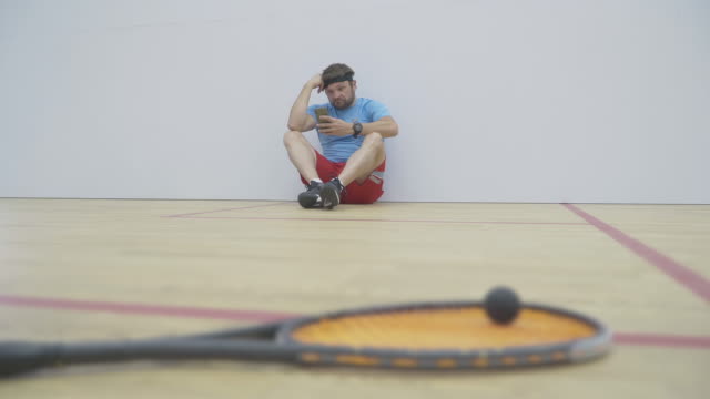 Wide shot of serious sportsman sitting against the wall in gym and using smartphone. Portrait of brunette bearded Caucasian man having break in training. Blurred racket and ball lying at front