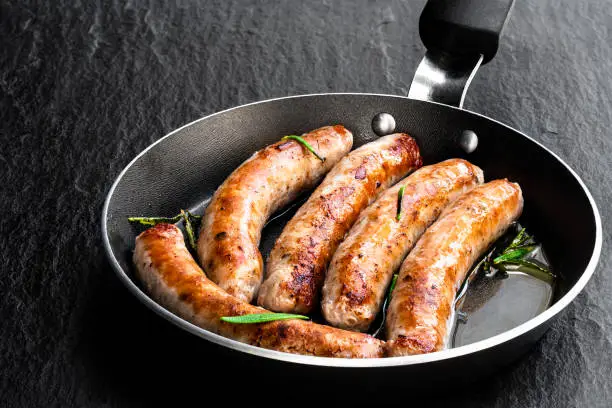 Photo of Delicious pork chipolatas sausages in a frying pan on black stone background